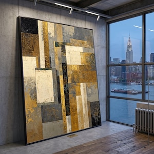 May include: Large abstract painting with a black frame. The artwork features a geometric design with gold, white, gray, and black rectangular shapes. The painting is displayed in a modern room with a city view through a large window.