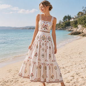 Op de afbeelding: Een witte mouwloze maxi-jurk met een vierkante halslijn en brede schouderbanden. De jurk is versierd met een herhalend verticaal patroon van kleurrijke bloemmotieven in rood, geel en groen, met een rok die uit verschillende lagen bestaat.
