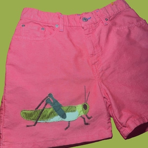 May include: Pink denim shorts with a grasshopper applique. The shorts have a classic five-pocket design and a button closure. The grasshopper is green, yellow, and blue.