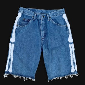 May include: Blue denim shorts featuring white fabric skeleton bone appliques running down each outer leg. The shorts have a classic five-pocket design, a button and zipper fly closure, and a raw, frayed hemline at the bottom of each leg.