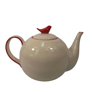 May include: A round, cream-colored ceramic teapot featuring a glossy finish. The teapot has a curved handle and a spout with red trim. A small, red, stylized bird figurine sits atop the center of the lid, serving as a decorative handle.