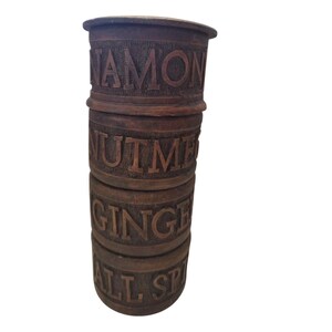 Puede incluir: Una pila vertical de cuatro recipientes cilíndricos de madera marrón para especias. Cada recipiente presenta letras talladas y hundidas con un fondo texturizado. Las etiquetas dicen CINNAMON, NUTMEG, GINGER y ALL SPICE de arriba a abajo.