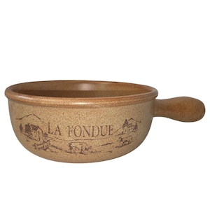 May include: A round, tan ceramic bowl with a single handle on the side. The exterior features a speckled texture and a dark brown printed illustration of a rural landscape with cows and the text LA FONDUE centered on the front of the vessel.