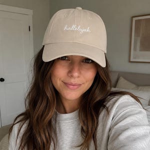 May include: A beige baseball cap with the word "hallelujah" embroidered in white script. The cap is made of a soft fabric and has a curved brim. The hat is a neutral color and can be worn with many outfits.