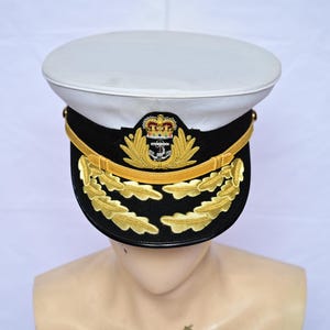 Könnte beinhalten: Eine Marineoffiziersmütze mit weißem Oberteil, schwarzem Band und goldenen Details. Die Mütze hat ein goldenes Emblem mit Krone und Anker sowie goldene blattähnliche Designs. Die Mütze wird auf einem Schaufensterkopf präsentiert.