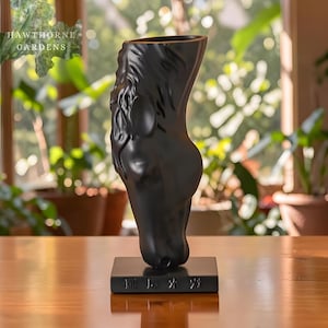 Pode incluir: Um vaso de cerâmica preta em forma de cabeça de cavalo, montado em uma base retangular. O vaso apresenta uma escultura detalhada da crina e das características faciais do cavalo. A base tem texto. O logotipo da Hawthorne Gardens está no canto superior esquerdo.
