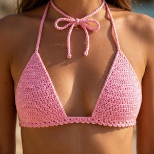 Puede incluir: Top de bikini de ganchillo rosa claro con cuello halter y detalle de lazo. Las copas triangulares están conectadas por una correa halter que se ata en la parte posterior del cuello. El borde inferior de las copas tiene un diseño festoneado.
