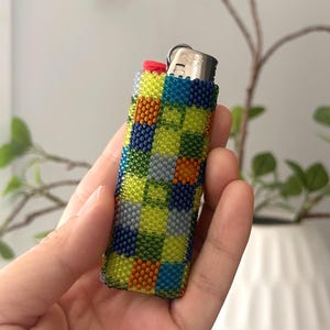 May include: A rectangular lighter encased in a beaded cover featuring a checkered pattern of lime green, navy blue, light gray, and orange glass beads. The lighter has a metallic silver top and a small red ignition button at the top corner.