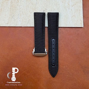 May include: A black watch strap made of a textured fabric material. The strap consists of two pieces, one featuring a silver-toned metal deployment clasp and the other with a series of punched holes for adjustment. The logo Customization STRAPS is visible.