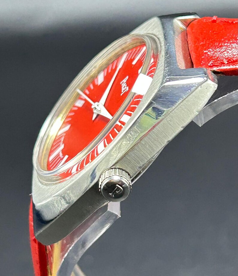 Puede incluir: Un reloj de pulsera con una esfera de color rojo vibrante y una caja de metal plateado pulido. El reloj cuenta con una correa de cuero rojo brillante y una peque&ntilde;a corona en el lateral grabada con la letra P. La esfera muestra el nombre Piaget en blanco.