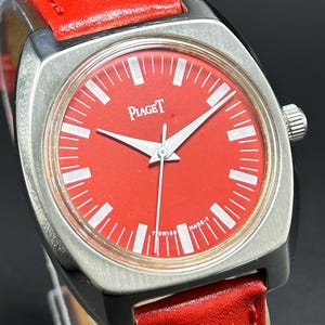 Puede incluir: Un reloj de pulsera con esfera roja brillante y caja de metal plateado cepillado. La esfera presenta marcadores de hora blancos, manecillas plateadas y el texto Piaget y Swiss Made-T. El reloj est&aacute; sujeto a una correa de cuero roja con costuras.