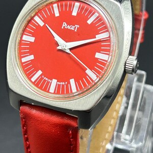 Puede incluir: Un reloj de pulsera Piaget con una esfera roja vibrante, marcadores de hora blancos y manecillas plateadas. El reloj tiene una caja cuadrada de color plateado cepillado con bordes redondeados y una correa de cuero roja con costuras.