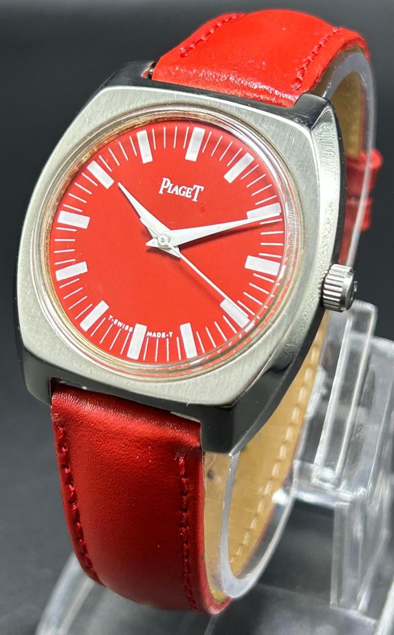 Puede incluir: Un reloj de pulsera Piaget con una esfera roja vibrante, marcadores de hora blancos y manecillas plateadas. El reloj tiene una caja cuadrada de color plateado cepillado con bordes redondeados y una correa de cuero roja con costuras.