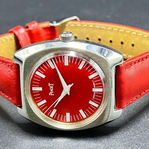 Puede incluir: Un reloj de pulsera con esfera roja brillante y caja de metal plateado. El reloj presenta marcadores de hora blancos, manecillas plateadas y el texto Piaget impreso en la esfera. Se sujeta con una correa de cuero roja con hebilla plateada.