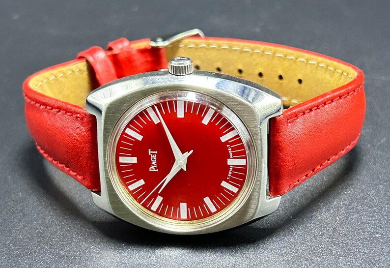 Puede incluir: Un reloj de pulsera con esfera roja brillante y caja de metal plateado. El reloj presenta marcadores de hora blancos, manecillas plateadas y el texto Piaget impreso en la esfera. Se sujeta con una correa de cuero roja con hebilla plateada.