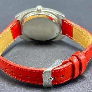 Puede incluir: Un reloj de pulsera con caja redonda de metal plateado y correa de cuero rojo vibrante. La correa incluye una hebilla de metal plateado, dos trabillas y un forro interior de color beige claro. La caja del reloj tiene una parte posterior plateada pulida.