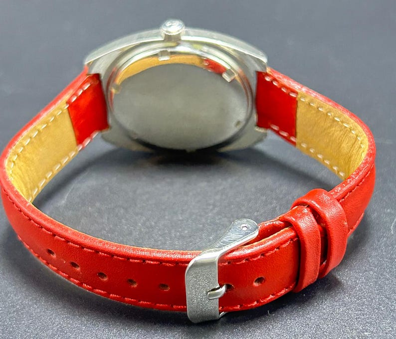 Puede incluir: Un reloj de pulsera con caja redonda de metal plateado y correa de cuero rojo vibrante. La correa incluye una hebilla de metal plateado, dos trabillas y un forro interior de color beige claro. La caja del reloj tiene una parte posterior plateada pulida.