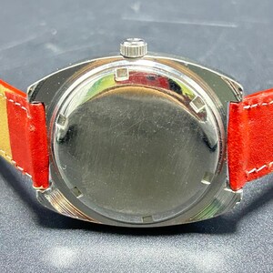 Puede incluir: Un reloj de pulsera con una caja circular de metal plateado y una parte posterior lisa y reflectante. El reloj cuenta con una peque&ntilde;a corona de cuerda en la parte superior y est&aacute; sujeto a una correa de cuero rojo vibrante con costuras claras.