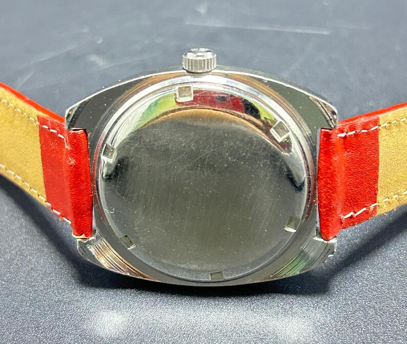 Puede incluir: Un reloj de pulsera con una caja circular de metal plateado y una parte posterior lisa y reflectante. El reloj cuenta con una peque&ntilde;a corona de cuerda en la parte superior y est&aacute; sujeto a una correa de cuero rojo vibrante con costuras claras.