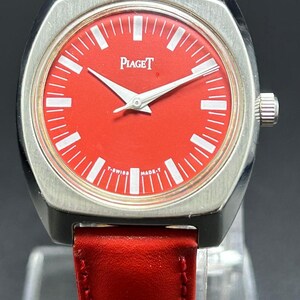 Puede incluir: Un reloj de pulsera Piaget con una esfera roja vibrante, marcadores de hora blancos y manecillas plateadas. El reloj tiene una caja cuadrada de tono plateado cepillado con bordes redondeados y una correa de cuero rojo lisa.