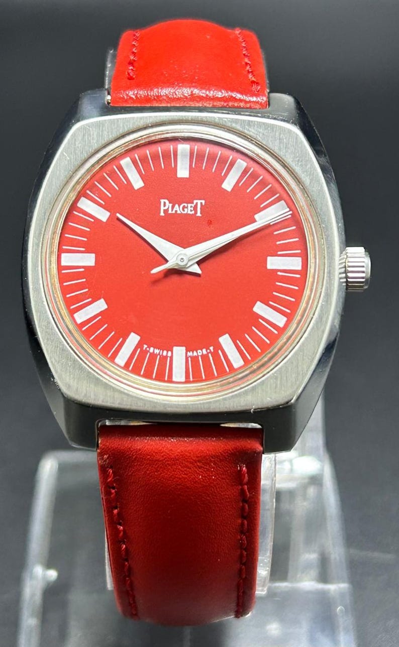 Puede incluir: Un reloj de pulsera Piaget con una esfera roja vibrante, marcadores de hora blancos y manecillas plateadas. El reloj tiene una caja cuadrada de tono plateado cepillado con bordes redondeados y una correa de cuero rojo lisa.