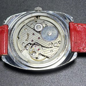 Puede incluir: Un reloj de pulsera plateado con la tapa trasera retirada para revelar el complejo movimiento mec&aacute;nico interior. El mecanismo est&aacute; grabado con el nombre de la marca Piaget y presenta engranajes visibles, un volante de inercia y un acabado texturizado. Una correa de cuero rojo brillante est&aacute; unida a la caja del reloj.