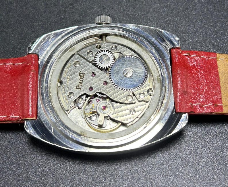 Puede incluir: Un reloj de pulsera plateado con la tapa trasera retirada para revelar el complejo movimiento mec&aacute;nico interior. El mecanismo est&aacute; grabado con el nombre de la marca Piaget y presenta engranajes visibles, un volante de inercia y un acabado texturizado. Una correa de cuero rojo brillante est&aacute; unida a la caja del reloj.