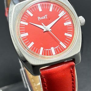 Puede incluir: Un reloj de pulsera Piaget con una esfera roja vibrante, marcadores de hora y manecillas en tono plateado. El reloj tiene una caja de metal plateado cepillado y una correa de cuero rojo liso. El texto Piaget aparece en la esfera, con T-SWISS MADE-T.