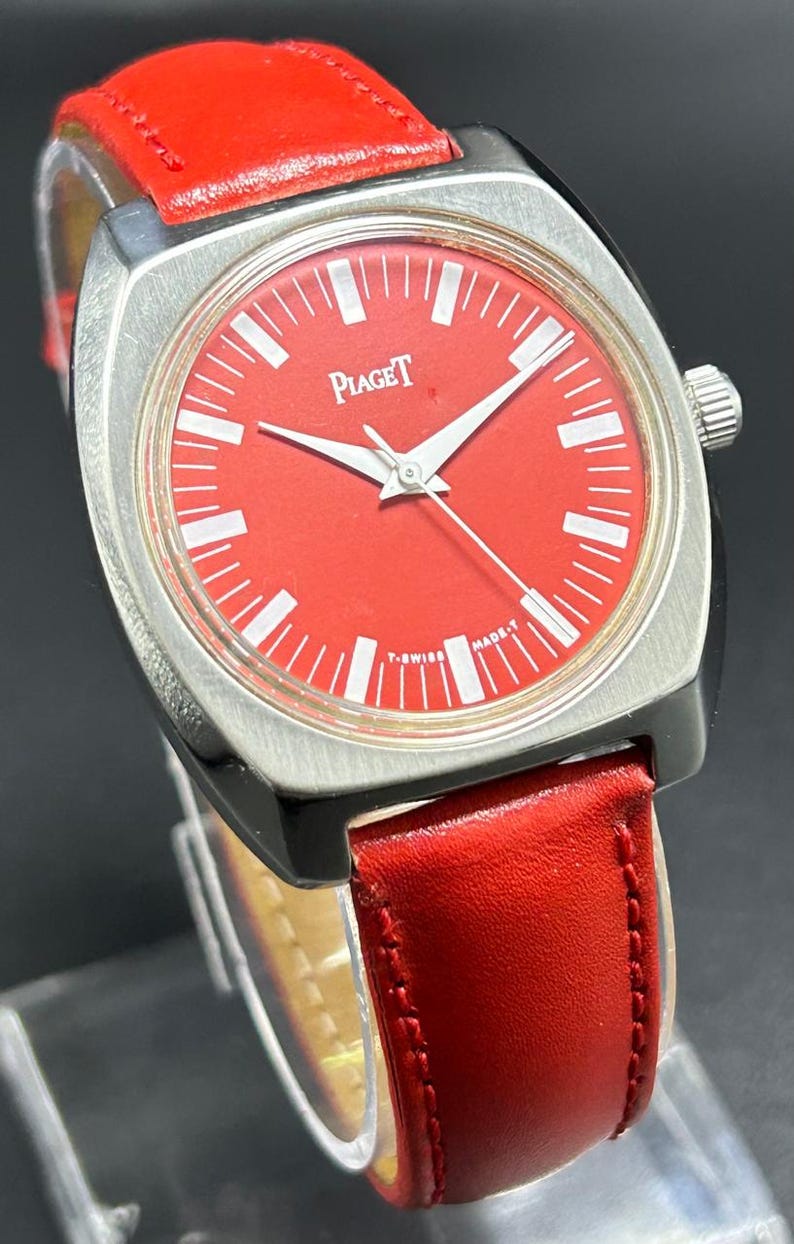 Puede incluir: Un reloj de pulsera Piaget con una esfera roja vibrante, marcadores de hora y manecillas en tono plateado. El reloj tiene una caja de metal plateado cepillado y una correa de cuero rojo liso. El texto Piaget aparece en la esfera, con T-SWISS MADE-T.