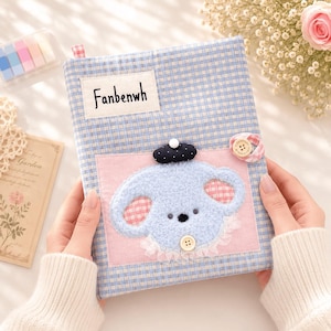 May include: A fabric-covered notebook featuring a light blue and white gingham pattern. It includes a white rectangular label with the text Fanbenwh, a pink square patch, and a fuzzy blue animal applique wearing a black beret and a white button.
