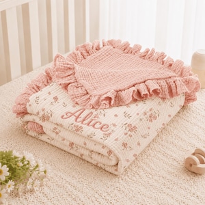 Puede incluir: Una manta de muselina doblada con base color crema, estampado floral rosa pequeño y borde con volantes rosa sólido. El nombre Alice está bordado en cursiva rosa en la parte frontal. La tela tiene una apariencia texturizada y arrugada.