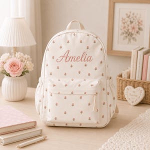 May include: A cream-coloured backpack featuring a repeating pattern of small pink rosebuds and green leaves. The name Amelia is embroidered in pink script on the front. The bag includes a top handle, adjustable straps, and a front zippered pocket.