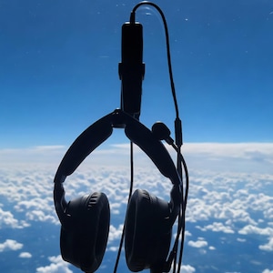 May include: Black aviation headset with a boom microphone, hanging against a backdrop of a bright blue sky and fluffy white clouds. The headset has large, round earcups and a flexible headband. The microphone is positioned near the left earcup.