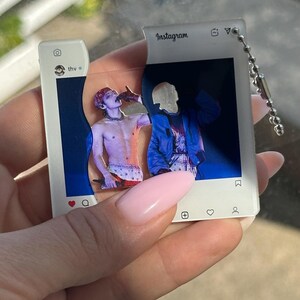 May include: A two-piece acrylic keychain shaped like an Instagram post interface. The white frame features the text Instagram, thv, and social media icons. The center displays a split image of a person in a blue jacket and a shirtless person.