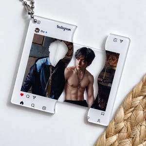 Puede incluir: Un llavero de acrílico transparente con forma de interfaz de publicación de Instagram y cadena de bolas. El diseño muestra una imagen dividida de una persona de pelo oscuro, un torso y una chaqueta oscura, con el texto Instagram y thv visible.