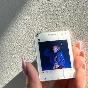 May include: A square white acrylic keychain featuring a printed Instagram interface design. The center displays a photograph of a person in a blue jacket. The top includes the text Instagram and thv, while the bottom shows standard navigation icons.