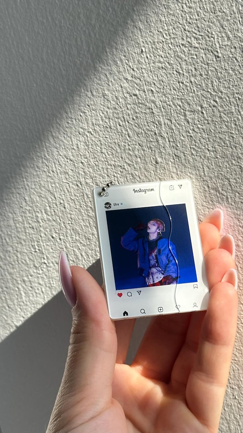 May include: A square white acrylic keychain featuring a printed Instagram interface design. The center displays a photograph of a person in a blue jacket. The top includes the text Instagram and thv, while the bottom shows standard navigation icons.
