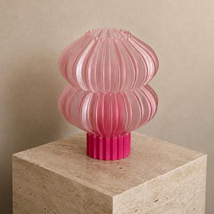 Può includere: Una lampada da tavolo caratterizzata da un paralume in vetro plissettato rosa chiaro, traslucido e a due livelli, con una silhouette arrotondata e bulbosa. La lampada è sostenuta da una solida base cilindrica magenta vivace con dettagli scanalati.