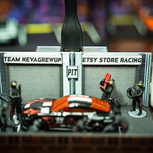 May include: A miniature racing pit stop diorama featuring a black and orange toy car, several small mechanic figurines, and grey garage structures. The central garage wall displays the text TEAM NEVAGREWUP, PIT, and ETSY STORE RACING in black lettering.