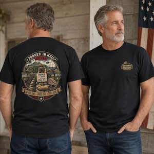 Op de afbeelding: Een zwart t-shirt met een afbeelding op de achterkant en voorkant. De achterkant toont een soldaat in een loopgraaf met de tekst FORGED IN HELL, VIETNAM, 1965-1973, HOME IS WHERE YOU DIG IT, en IN THE DIRT UNDER FIRE STILL HERE. De voorkant heeft een klein logo.