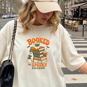 Puede incluir: Una camiseta de color marfil con un gráfico de tres fantasmas leyendo libros apilados unos sobre otros, con el texto "BOOKED FOR SPOOKY SEASON" en naranja y verde. La camiseta se combina con una gorra de béisbol beige y un bolso negro.