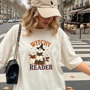 Puede incluir: Una camiseta color marfil con un gráfico de tres fantasmas leyendo libros y el texto "WITCHY READER" en marrón y naranja. Se combina con una gorra de béisbol beige con un pequeño logotipo oscuro y un bolso de hombro negro con cadena.