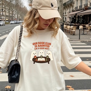 Puede incluir: Una camiseta de color marfil con un gráfico de tres fantasmas sentados a una mesa redonda con un libro abierto y tazas. El texto dice: OUR BOOK CLUB IS HAUNTED (IN A GOOD WAY). La camiseta se combina con una gorra beige y un bolso negro.