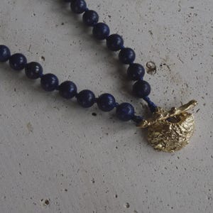 Könnte beinhalten: Eine Halskette mit einer Reihe dunkelblauer, kugelförmiger Perlen, die auf einer passenden blauen Schnur aufgereiht sind. Die Kette wird mit einem goldfarbenen Knebelverschluss geschlossen, der eine texturierte, unebene Oberfläche aufweist.