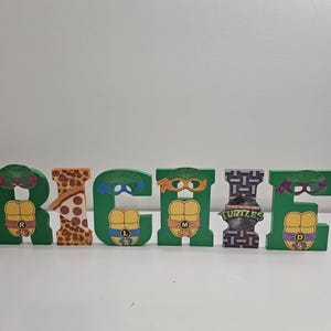 Può includere: Lettere di legno che compongono il nome "RICHIE" a tema Tartarughe Ninja. Ogni lettera è verde con una maschera di tartaruga e un design a guscio. La lettera "I" è un design grigio, a motivi, con le parole "Teenage Mutant Ninja Turtles".