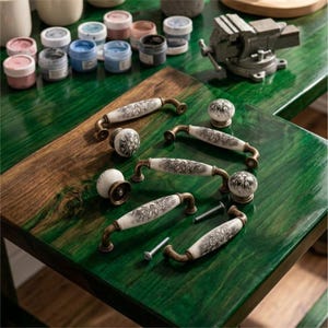 May include: A collection of ceramic drawer pulls and knobs featuring a white base with intricate black floral patterns and antique brass-toned metal hardware. The set includes several arched handles and round knobs, accompanied by small metal mounting screws.