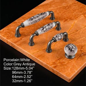 Puede incluir: Tres tiradores de armario y un pomo redondo con centros de porcelana blanca con patrones florales grises y herrajes de metal gris antiguo. Los tamaños son 128 mm, 96 mm, 64 mm y 32 mm para el pomo redondo.