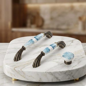 May include: Two arched cabinet pulls and one round cabinet knob featuring white ceramic centers with blue cross-hatched patterns and antique bronze-toned metal ends with a ribbed, fluted design on the mounting bases.