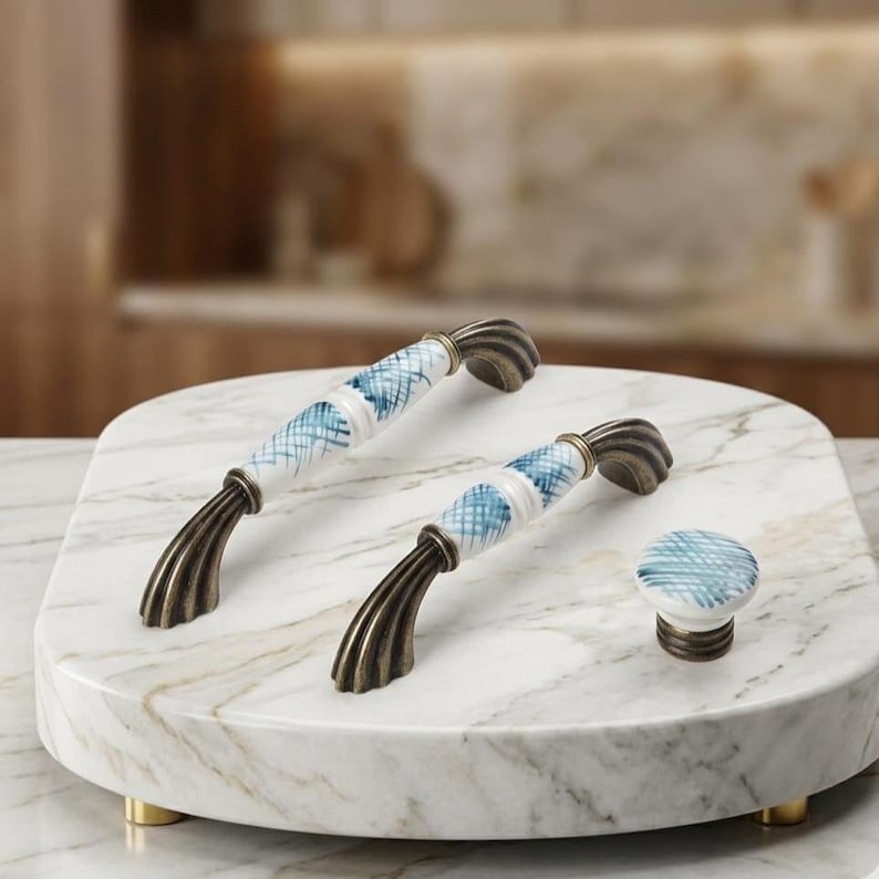 May include: Two arched cabinet pulls and one round cabinet knob featuring white ceramic centers with blue cross-hatched patterns and antique bronze-toned metal ends with a ribbed, fluted design on the mounting bases.