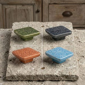 May include: Four square cabinet knobs with embossed botanical leaf patterns. The set includes one green, one dark grey, one terracotta orange, and one light blue knob, each featuring a flat top surface and a tapered base for mounting.